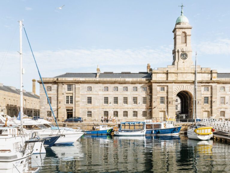 A group of small boats bob in the harbour at Royal William Dock Yard, the Plymouth office building in the backdrop, mirrored in the water's surface.