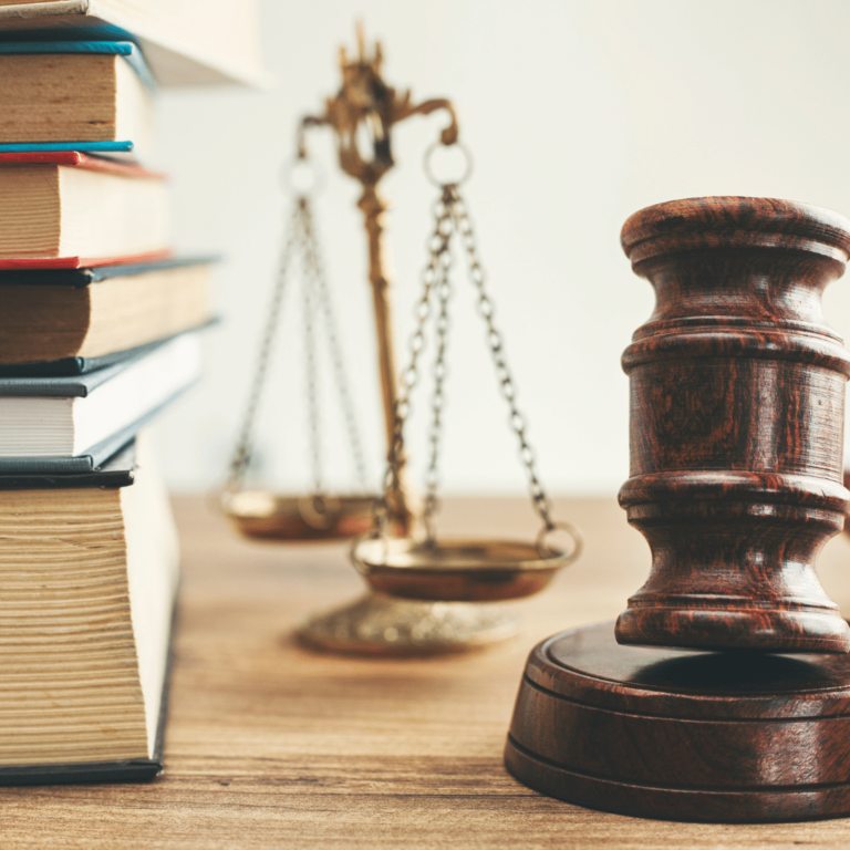 A gavel rests on its block next to a tower of books in the foreground, behind it is a set of brass scales.