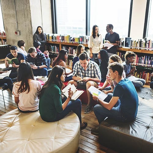 A large group of students sit on low leather seating while they study in a library.