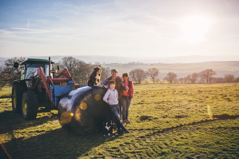 A family of farmers, accompanied by their two border collies, pose as they lean against a hay bale, which is propped up by a tractor, as the sun sets over their property.