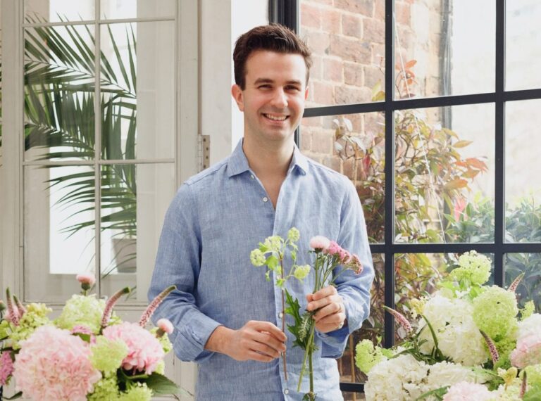 Bloom & Wild co founder and CEO Aron Gelbard stands in a greenhouse holding flowers while grinning at the camera.