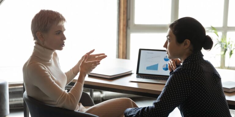 Two female office workers sit in front of a computer engaged in a passionate discussion.