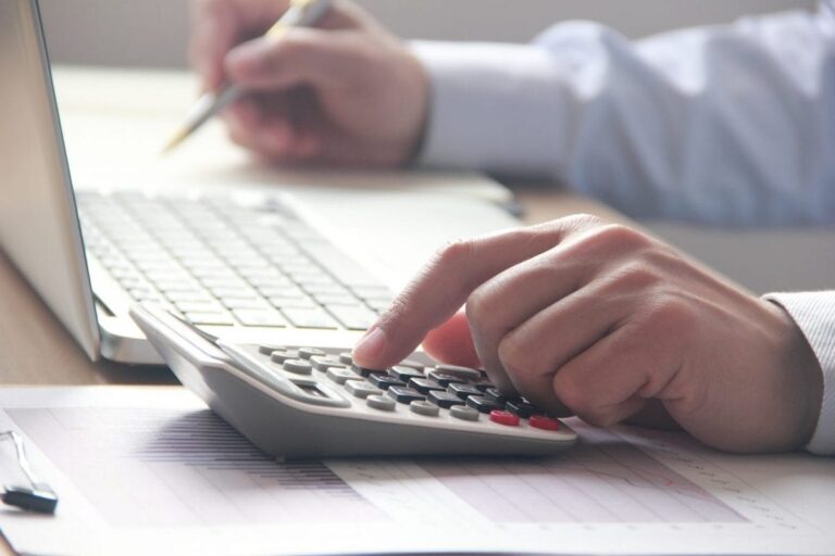 A male office worker sits in front of a lap top computer and enters figures into a calculator.