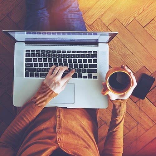 An aerial view of a person sitting on a wooden floor, holding a coffee cup, and working on their laptop computer.