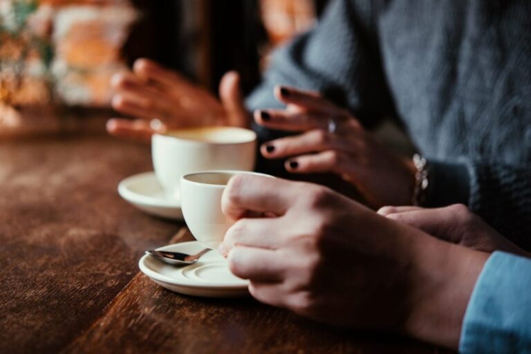 Two coffee cups resting on a table while two friends converse in a coffee shop.