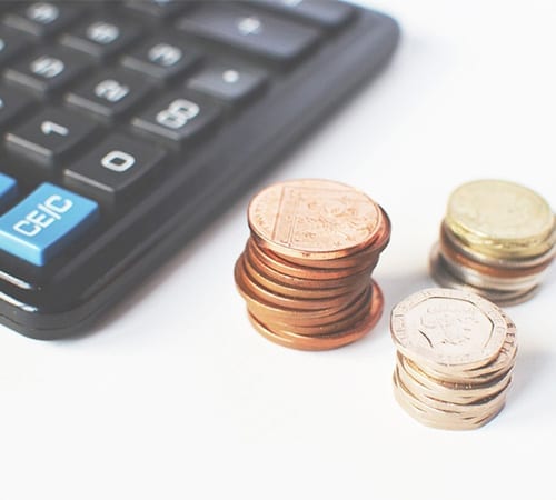 Three piles of various coins stand beside a calculator with a crisp white backdrop.