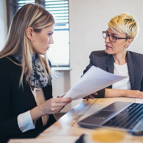 Two women in suits are seated at a computer, engaged in a serious discussion.