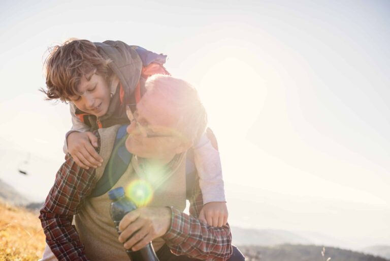 A grandfather and his grandson look lovingly at each other as the young boy rides on his grandfather's back, setting out across a field, on an adventure.