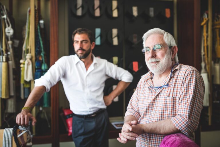 A father and his adult son are standing, leaning on chairs, at their family-owned upholstery business.