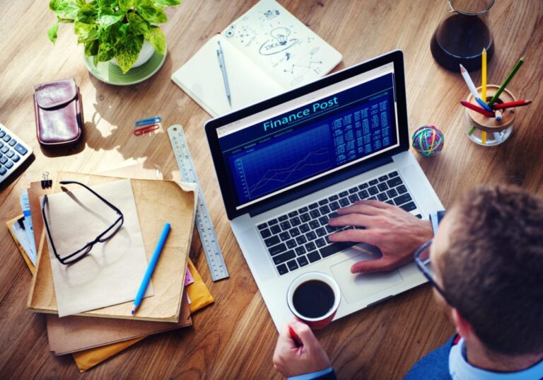 A man is looking at the Finance Post on his laptop situated on a desk which has papers and pens scattered over it.