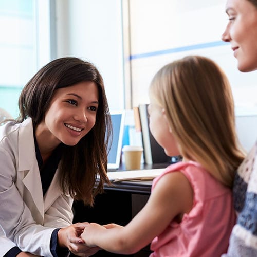 A female GP smiles as she leans forward and holds the hands of a child patient.