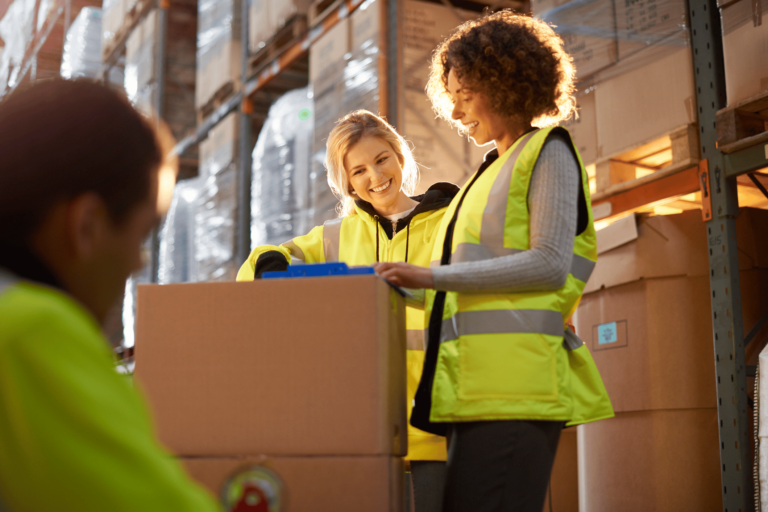 Two female warehouse operatives in hi-vis vests surrounded by cardboard boxes are signing off paperwork.