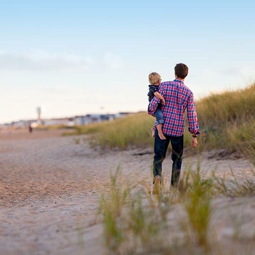 A father strolls down the beach holding his son on one hip.