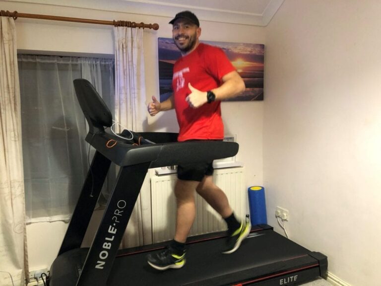 Jason Mitchel is smiling and giving a double thumbs-up to the camera as he runs on his treadmill at home.