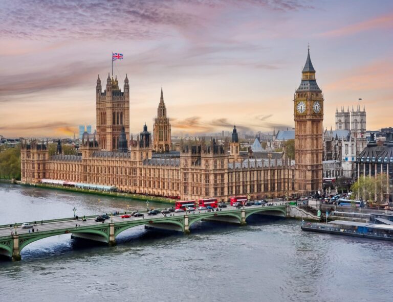 A view across the Thames river towards Big Ben and the Houses of Parliament, the Union Jack is raised high against a sky of purple hues.