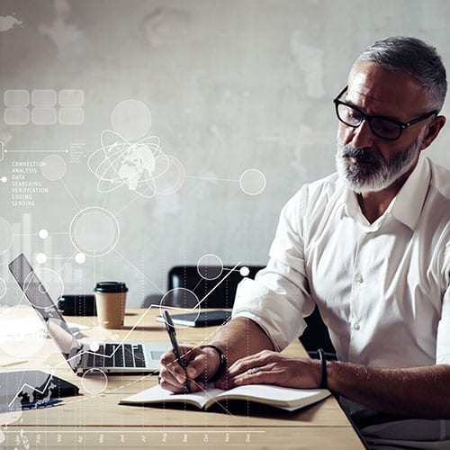 A man in glasses sits at a desk entering tax calculations into a note pad while his laptop sits open.