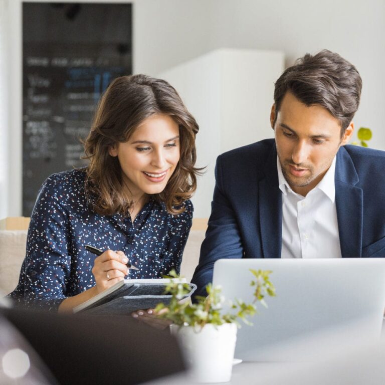 A man and woman lean in to look at a laptop screen while the woman makes notes on a paper pad.