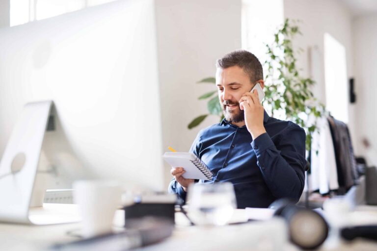 An office worker sits in front of a computer whilst looking at a notepad with his mobile phone held to his ear.