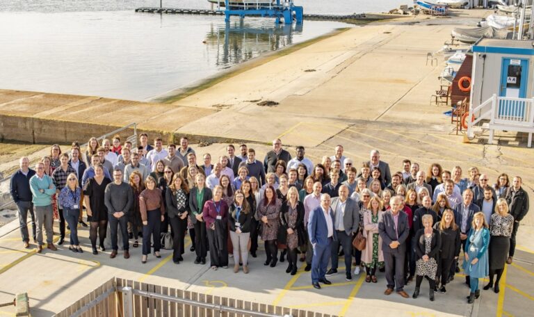 An overhead view captures a large assembly of Francis Clark employees at a dockyard, all smiles directed upwards at the camera, in celebration of their recognition as a great workplace.