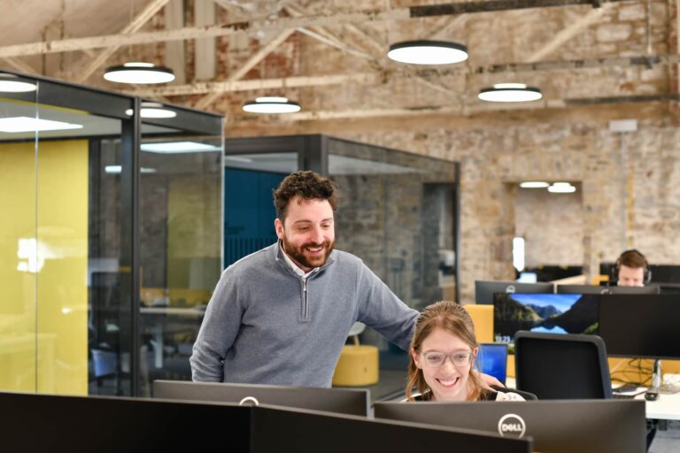 Archie Willmot leans on the back of his colleague, Lily Gammon's chair, as they both smile at her computer screen in the Plymouth office.