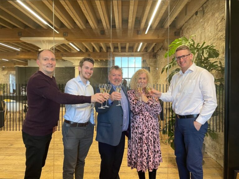 Five individuals are celebrating a company acquisition with a wine toast in a heritage-style building.