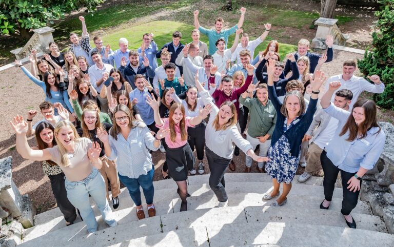 A large group of new trainees pose on steps, hands raised in celebration.