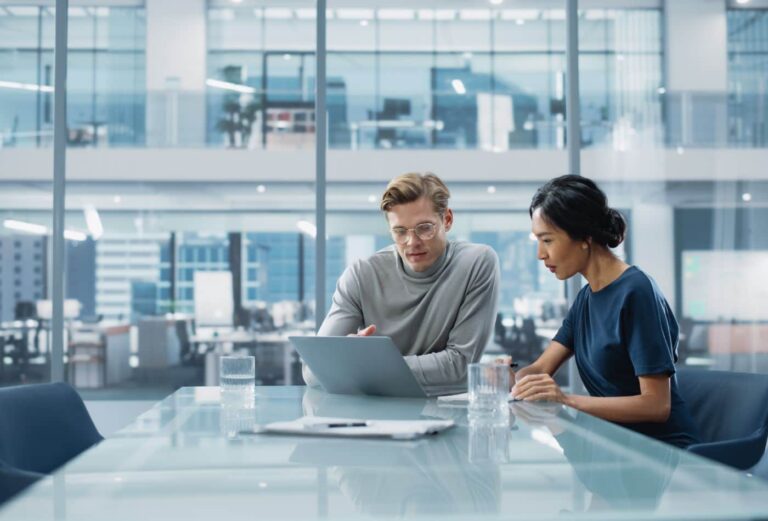 A male and female colleague look down at a laptop screen while sitting in a modern glass walled office.