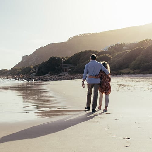 An older couple embraces each other while walking on the beach, enjoying the sunset.