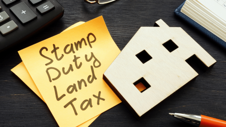 A wooden house cut out lies on a desk, on top of a Post-it note inscribed in black marker with 'Stamp Duty Tax' .