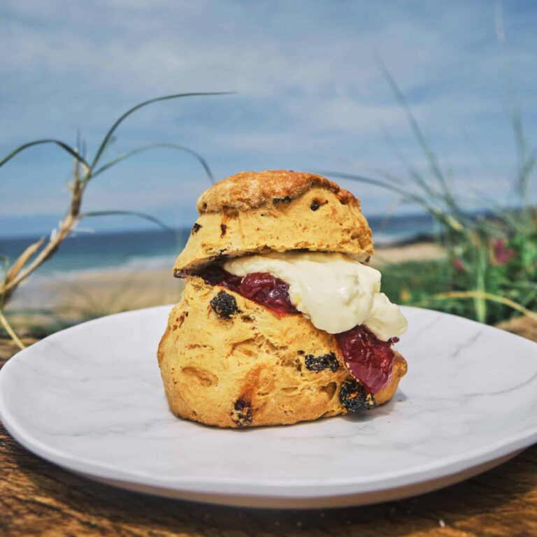 A current scone with jam and cream sits on a plate with the beach in the background.