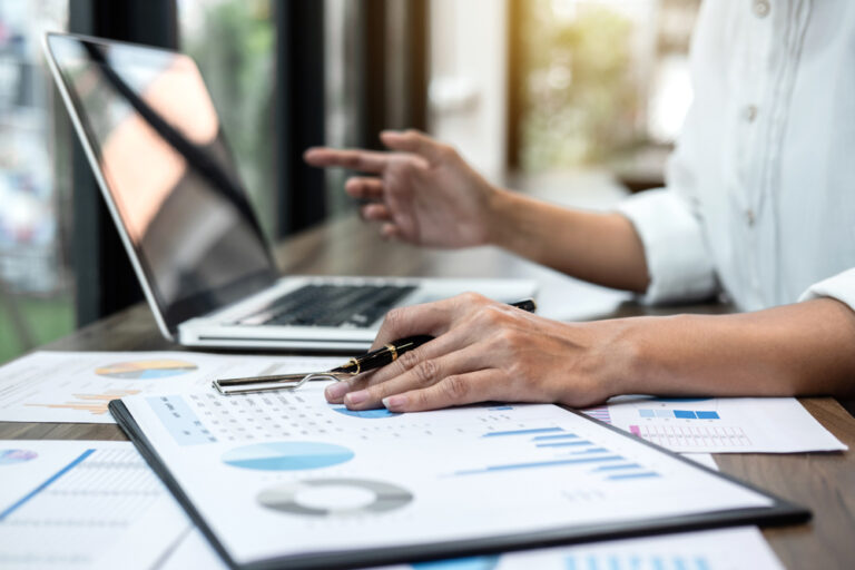 A calendar and a laptop are positioned on a desk, with an individual holding a pen in one hand while pointing at the laptop screen with the other.