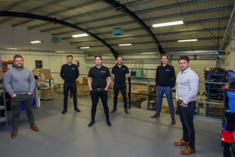 The iOutlet team stands, hands in their pockets, in a hangar filled with boxes.