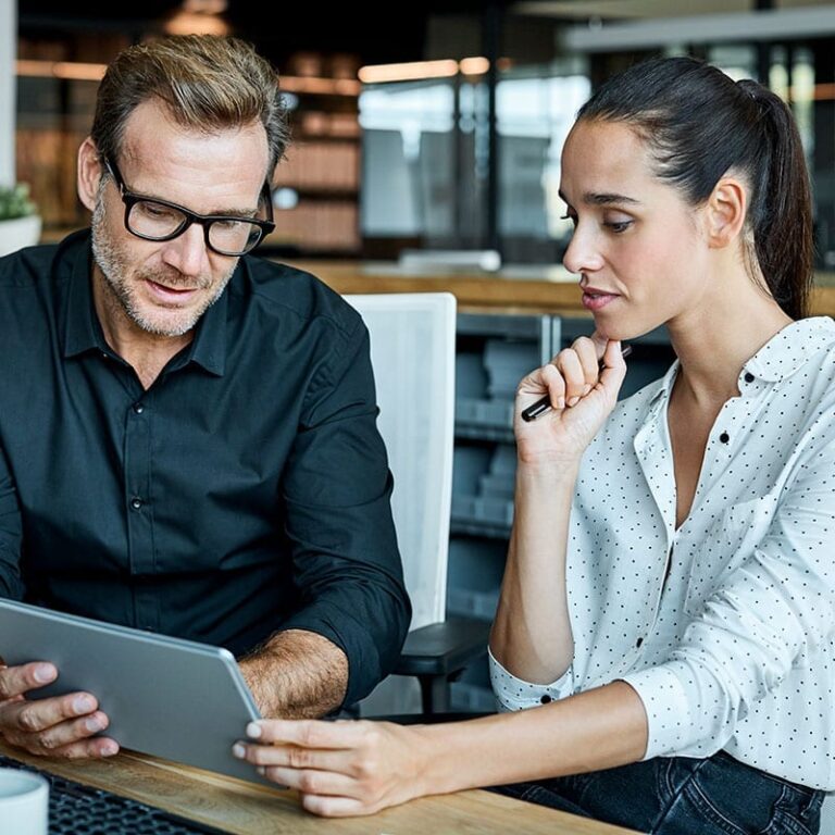 A male and female co-worker sit in an office looking at a computer tablet.