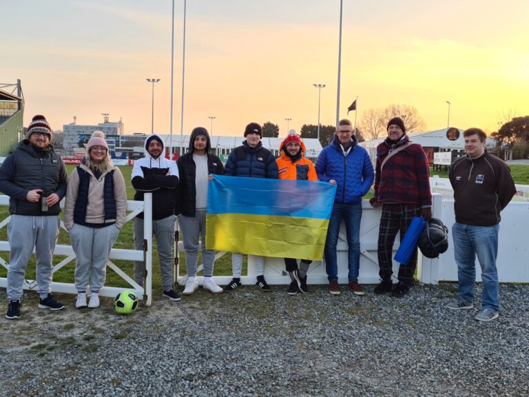 A group dressed in winter clothing stands beside a football pitch, holding a Ukrainian flag, after a fundraising event.