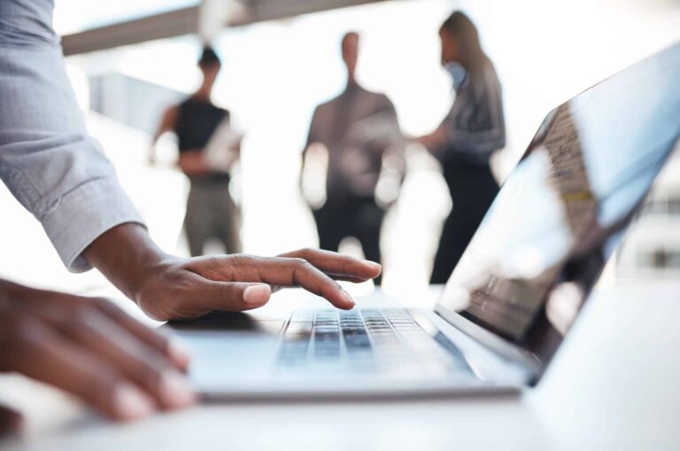 A close up picture two hands poised over a laptop with the blurred image of three colleagues stood in the background.