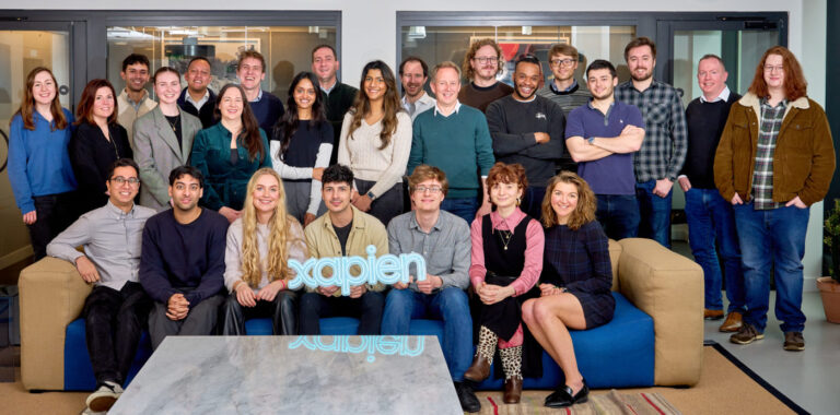 Some Xapien employees sitting on a sofa whilst others are standing, in their office smiling at the camera.