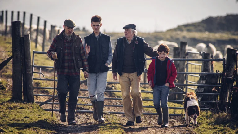 Three farmers, a boy, and a dog are walking in front of a metal gate that encloses cattle in a field.