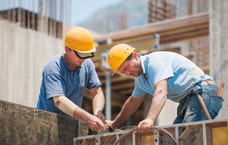 Two workmen in hardhats work together to feed copper wire through a hole at a construction site.