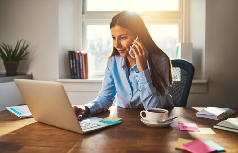 A business woman talks on her mobile phone whilst working at her computer at home.