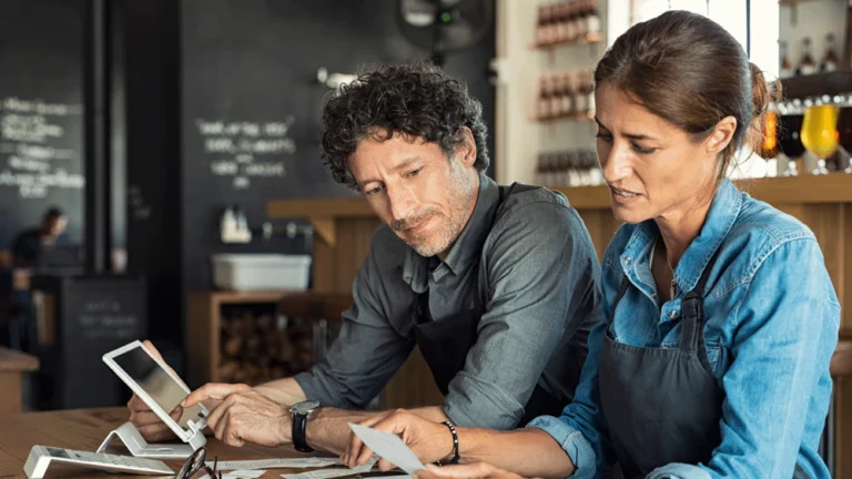 Two café owners check their receipts, while entering figures into an computer tablet.