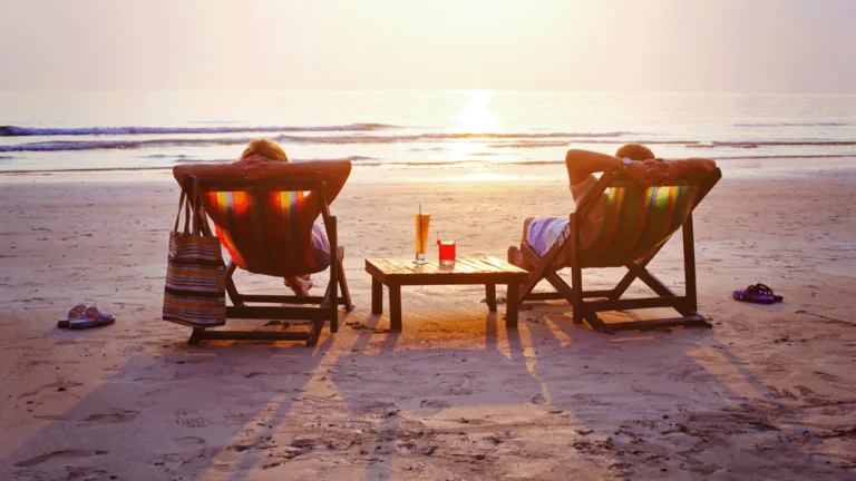 A couple are relaxing in deckchairs on a beach at sunset.