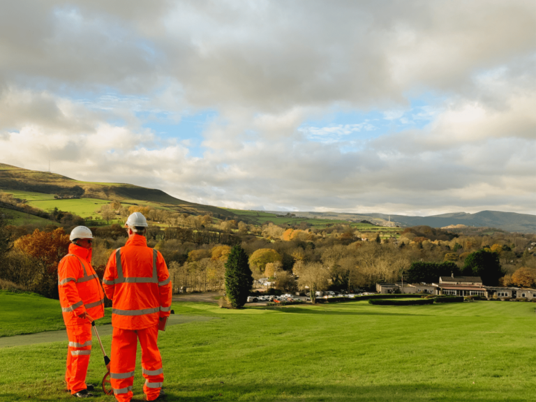 Two men in orange reflective overalls and hard hats standing talking in a field surrounded by trees with a building in car park in the distance.