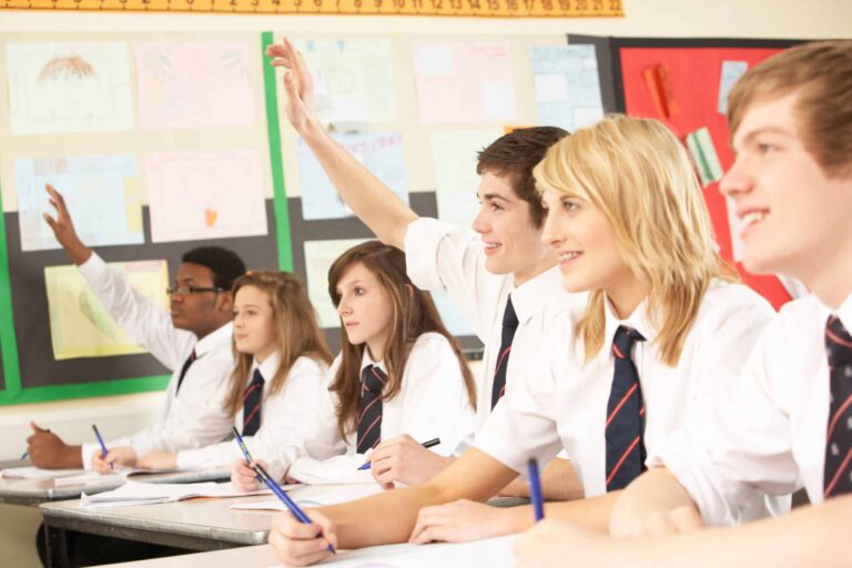 Six secondary school students sitting in class whilst two students have their hands up to answer a question.