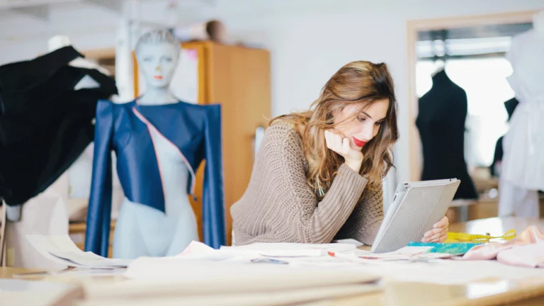 A female fashion designer surrounded by material and drawings looking at her i-pad