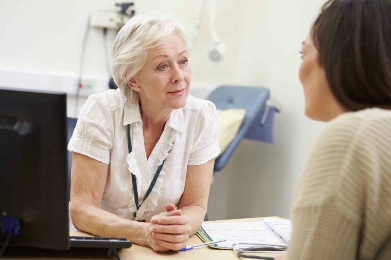 A female GP leans on her desk, hands clasped together, as she talks to her female patient.