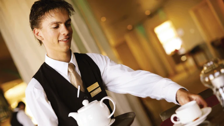 Waiter placing a cup on a table whilst carrying a tray with a tea pot on