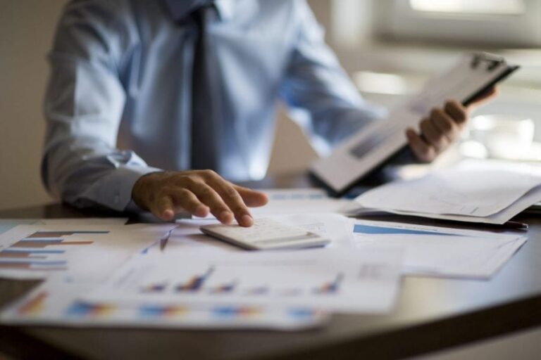 A male office worker sits at a desk surrounded by paperwork entering figures into his calculator.