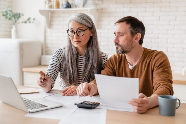 A couple sit at their kitchen table and make calculations while looking at paperwork and a laptop.