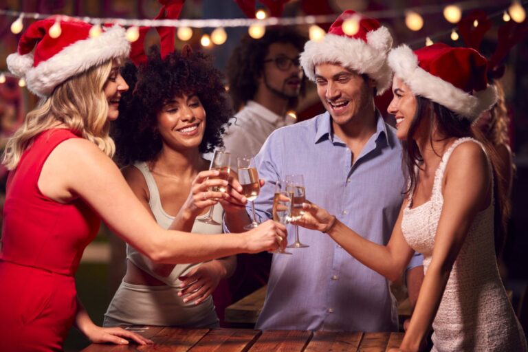 A group of young colleagues wearing Christmas hats stand in a circle and raise their glasses to make a toast.