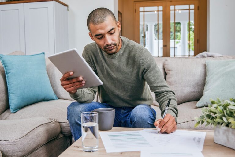 A man is seated on his sofa, a computer tablet in one hand, as he leans forward to fill in paperwork on the coffee table with his other hand.